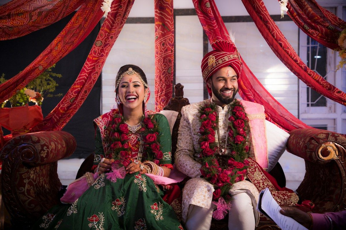 A smiling bride and groom in garlands seated together on a stage
